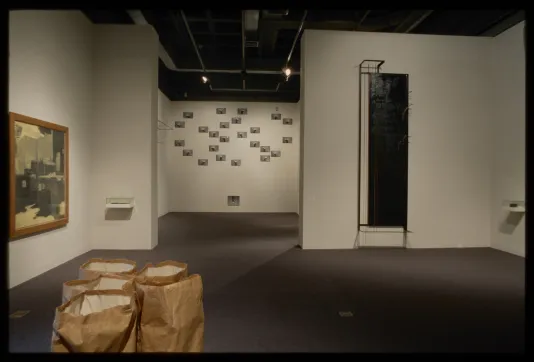 Paper sacks sit in gallery floor, photographs, small objects, and mixed media works of varying sizes hang on the walls.