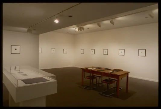 Table and glass case sit in front of nine small framed drawings of white background with tiny colored shape in the middle.