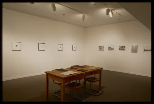 Wooden table with chairs and books sits in front of four drawings of small shape hung adjacent to five landscape photos.