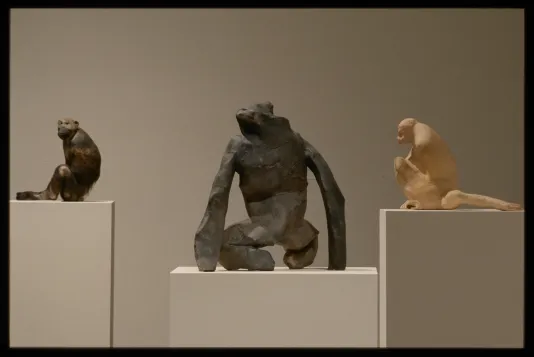 Three abstracted clay sculptures of apes sit on white gallery pedestals. From left to right: Brown and black, black and grey, tan. 