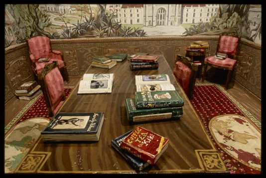 Colonial-style room with landscape wallpaper, wooden table with eleven colorful books, red cushioned chairs, and map carpeting. 