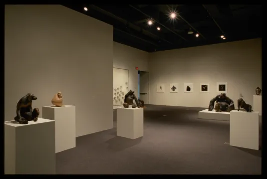 Gallery view of seven clay ape sculptures on pedestals, paper wall mural of text and apes heads, and five small ape paintings.