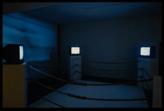 A boxing ring where the corners are made from pedestals with monitors. The blue light from the monitors shine on the walls.