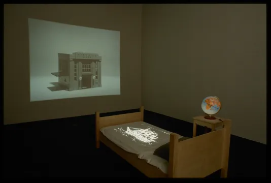 A bed and side table are set in front of a projection on the wall. A projection also shines on the bedsheets.