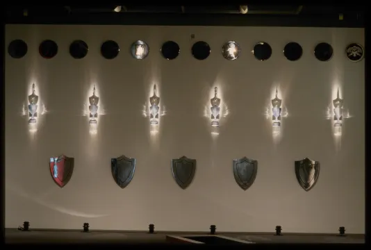 12 disks line the top of the gallery wall. Beneath them are six shining trophies. Lower in the wall are five shields.