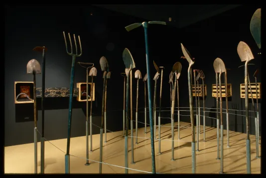 Various farming tools stand upright on a platform in the gallery. Each one casts a long shadow on the floor. 
