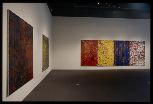 Three large abstract paintings covered entirely in colorful smears and splatters hang on adjacent gallery walls.