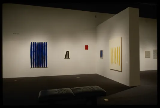 Hanging on gallery wall are five artworks of various sizes, with thick rounded lines of blue, black, red, blue, and yellow.