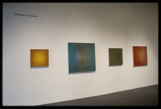 Four paintings of various sizes with solid colors of yellow, blue, green, and red which fade lighter in the middle hang on wall.