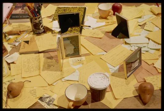 A tabletop littered with stationary, cups, gold frames, and fruit creates an overall image of muted yellow.