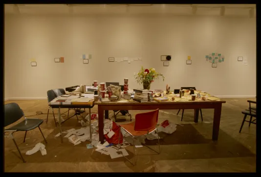 Two tables abut, littered with cups, books, papers, and a messy heap of paper below. Groupings of notes on the wall behind.