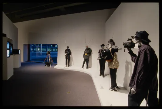 Five mannequins dressed in casual clothes and posed with surveillance gear line wall across from four tvs playing videos. 