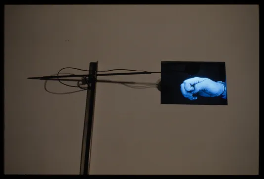 Art installation of metal and wired surveillance equipment connected to screen with close up of blue hand in shape of a flat fist.