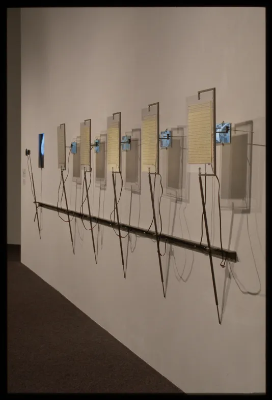Art installation of six metal and wired surveillance gear mounted on wall holding transcripts with videos playing behind them.