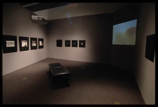 Nine black and white photos in black frames hang on adjacent walls. Bench sits in middle of room facing large screen playing video.