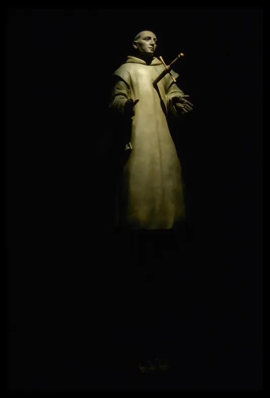 Spotlit sculpture in dark gallery room of polychromed bald saint in robes with hands outstretched and sword in chest.
