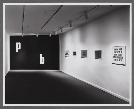 Black and white image of a room with p and b in white on a black wall. 5 framed paintings of text hang on a wall on the right.
