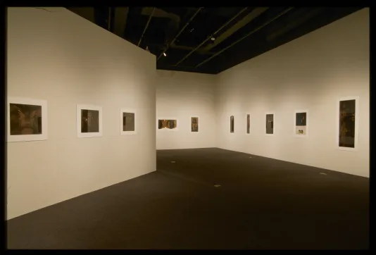 Sepia toned photographic prints, bordered in white, hang on the walls of a gallery space.