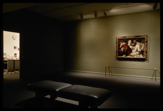 Dark gallery room with bench facing lit Caravaggio painting, off to side is doorway showing lit room with desk and documents.