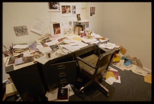 Desk and chair with disarray of art historian documents and office items covering the desk and spilling onto the floor.