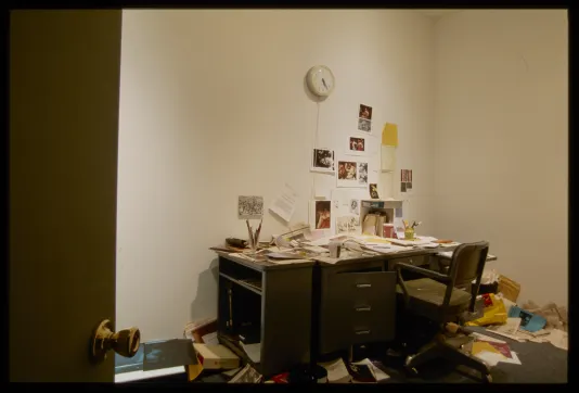 Door open revealing desk with art historian documents and office items in disarray covering desk and some wall and floor.