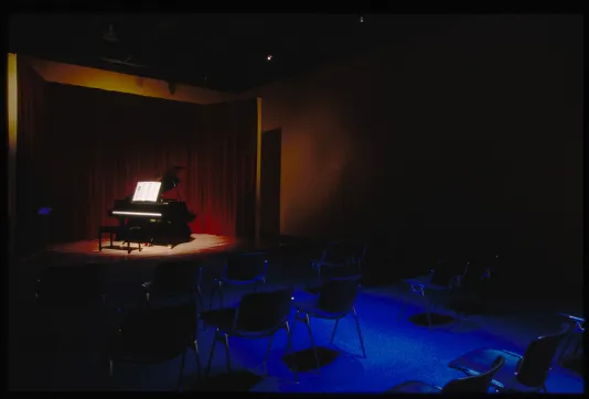 Dark chairs on blue floor set up for audience to view a spot-lit, black, grand piano before a red curtain on stage