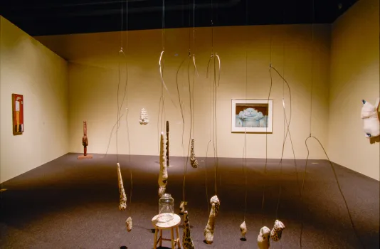 Organically-shaped objects hang from metal wires above a glass cloche in a gallery with sculptures, painting and a photograph.