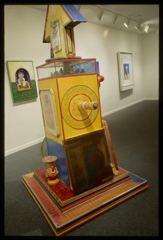 A large bright yellow sculpture sits on the forgeround of a gallery space.