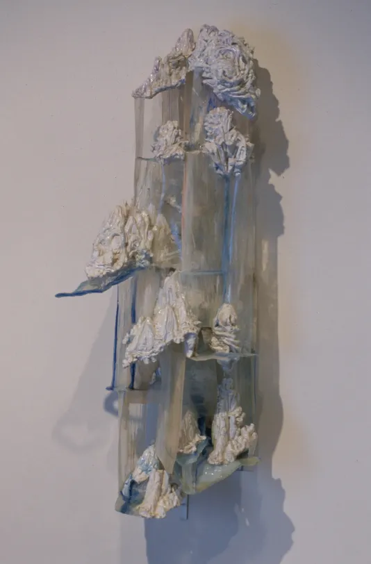Gray-cream-color organic rock-like forms cling to the edges of a clear jagged column in this sculpture hung on a wall.