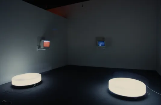 2 round discs of white light sit on the floor before 2 gray, wall-mounted monitors showing color images in a dark gallery.