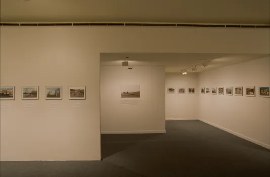 A row of framed color photographs hangs at eye level on white walls around 3 rooms of the gallery.