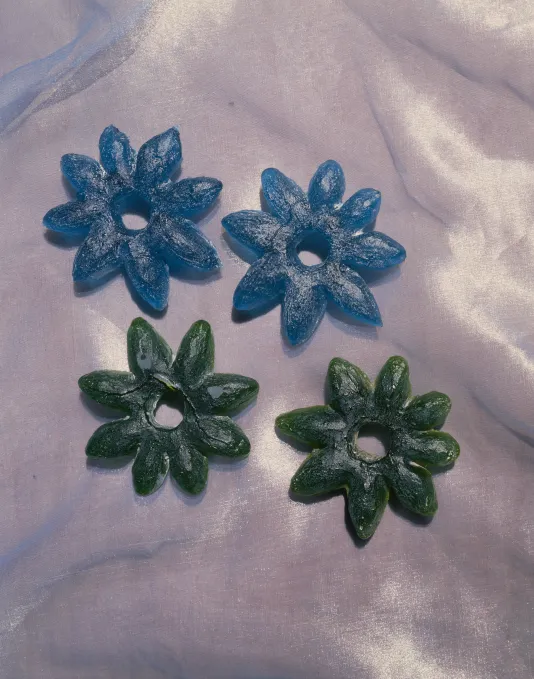 Blue, cast-glass stars, flowers and trivets surround an iridescent, blue-green disc on shiny, tan and cream organza fabric.