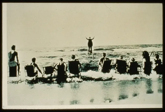 Seen from behind, a man in a tailcoat faces the horizon and conducts the waves as a row of sunbathers in beach chairs watch.