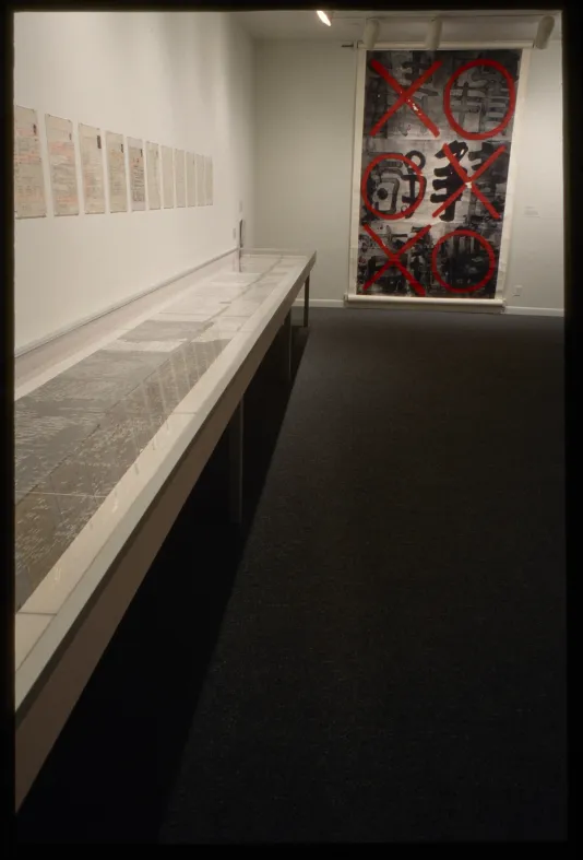 Small pieces hang above a long vitrine, and lead to a large scroll with red X’s and O’s covering calligraphic writing.