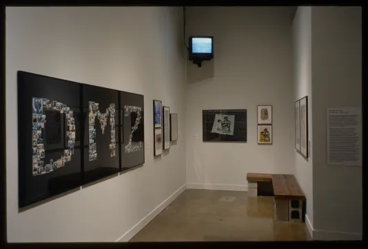 Hanging works include a piece with the collaged letters “D M Z”, a video plays on the far wall, a wood bench sits on blocks.