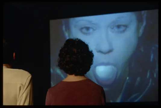 Two viewers watching a video with a young woman sticking out her tounge on the screen. 