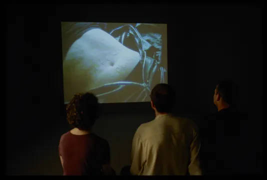 Three viewers sitting in a darkened gallery with an image of a male torso on the screen. 