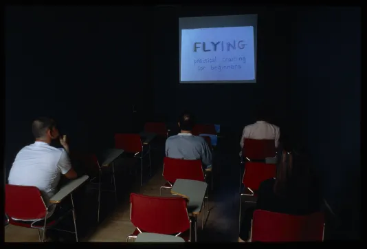 4 viewers sitting in a dark room in red chairs looking at a fim with the title Flying. 