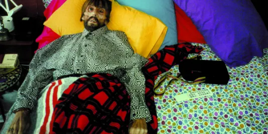  Photograph of bearded man with a sunken face on the edge of a bed covered in vibrant colored pillows and patterned sheets