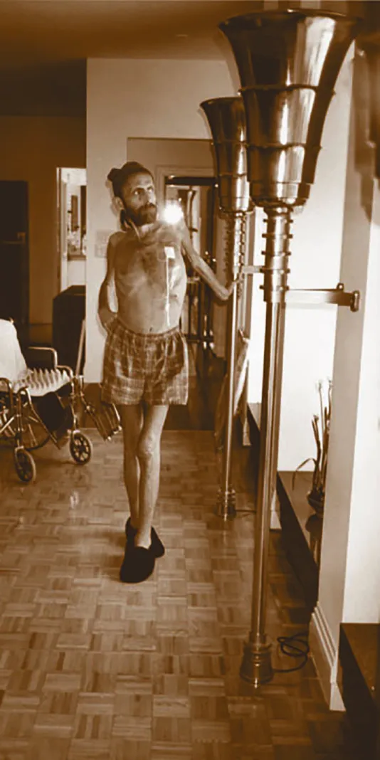 Sepia photograph of emaciated man standing in plaid boxers and slippers beside a wheelchair, gazing out the window