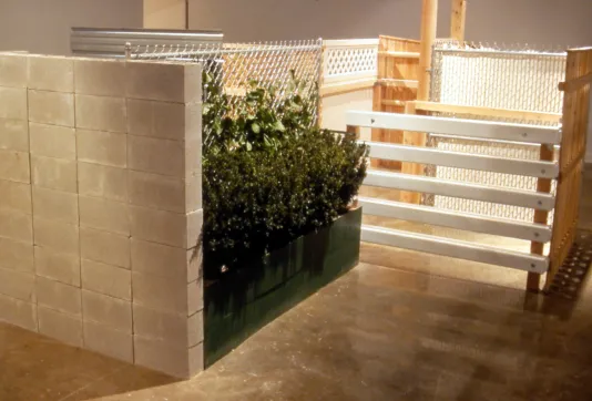 A row of green plants sits tucked behind a concrete colored wall, beside a series of metal, plastic, and wood fences.