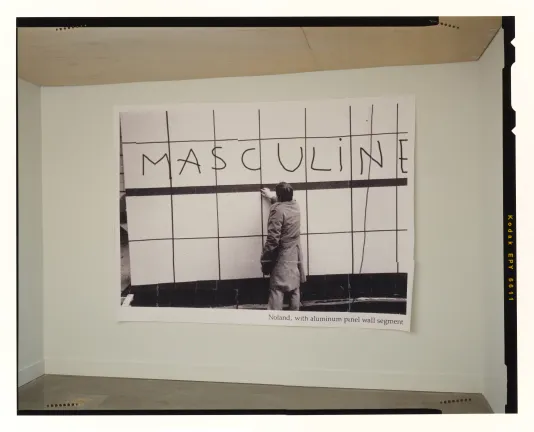 A black and white print of a man reaching out to touch a gridded aluminum wall. The word MASCULINE is handwritten above him. 
