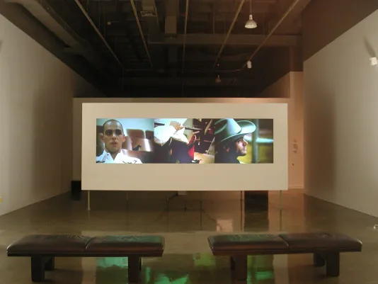 In a dimly lit space, a film screen displays 3 different closeup images of men in cowboy attire.