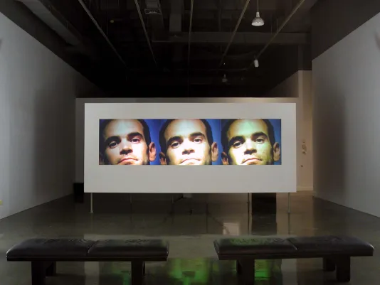 In a dimly lit space, a film screen displays 3 images of a man’s face, each image slightly different in coloration.