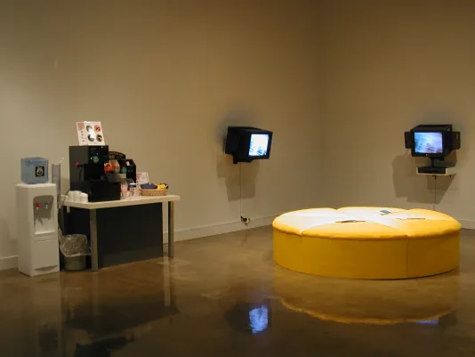 Pamphlets sit on a large round yellow ottoman, in a room with active wall mounted monitors, and a coffee and a water station.
