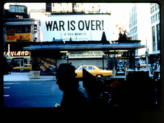 On the street, yellow taxis and people walking below a billboard in Times Square which reads “WAR IS OVER! IF YOU WANT IT!” 