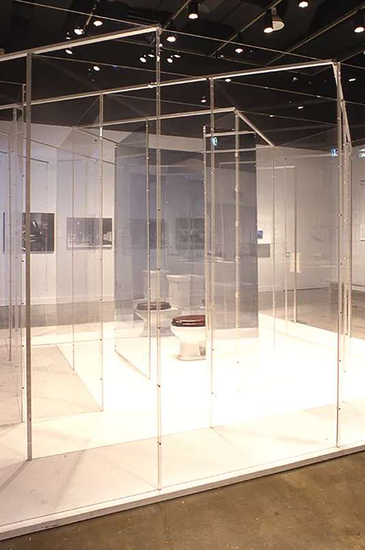 A white toilet with a brown lid sits amongst glass dividers in gallery space.