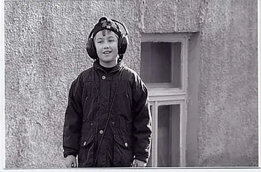 Black and white video still of a young boy wearing headphones over his ears, in front of a concrete building 