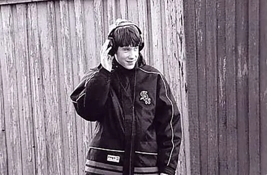 Black and white video still shows a teen boy with headphones over his ears, in front of a wooden fence