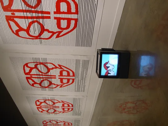 Covering the length of the gallery wall is a repeating image of a large red face-like design. A box monitor sits on the ground
